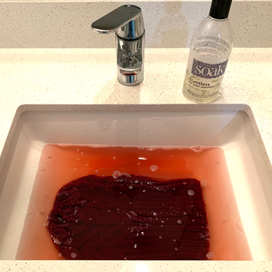 Sink full of water tinged with red dye from a knitted hat and a bottle of Scentless Soak