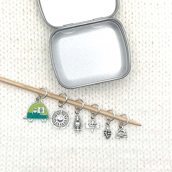 Set of one two-toned green and silver enamel tiny camper and five antiqued silver camping themed stitch markers on bamboo knitting needle with storage tin on white knitted background for knitting by Pretty Warm Designs