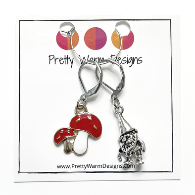 One red and white enamel toadstool crochet stitch marker and one antiqued silver gnome crochet stitch marker attached to silver ring hanging from white cardstock printed with Pretty Warm Designs logo and website URL