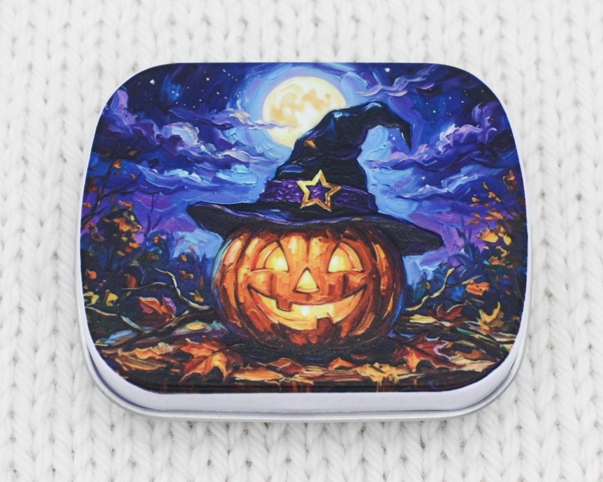 A small storage tin tfeaturing a Halloween illustration of a glowing jack-o'-lantern wearing a witch hat under a full moon