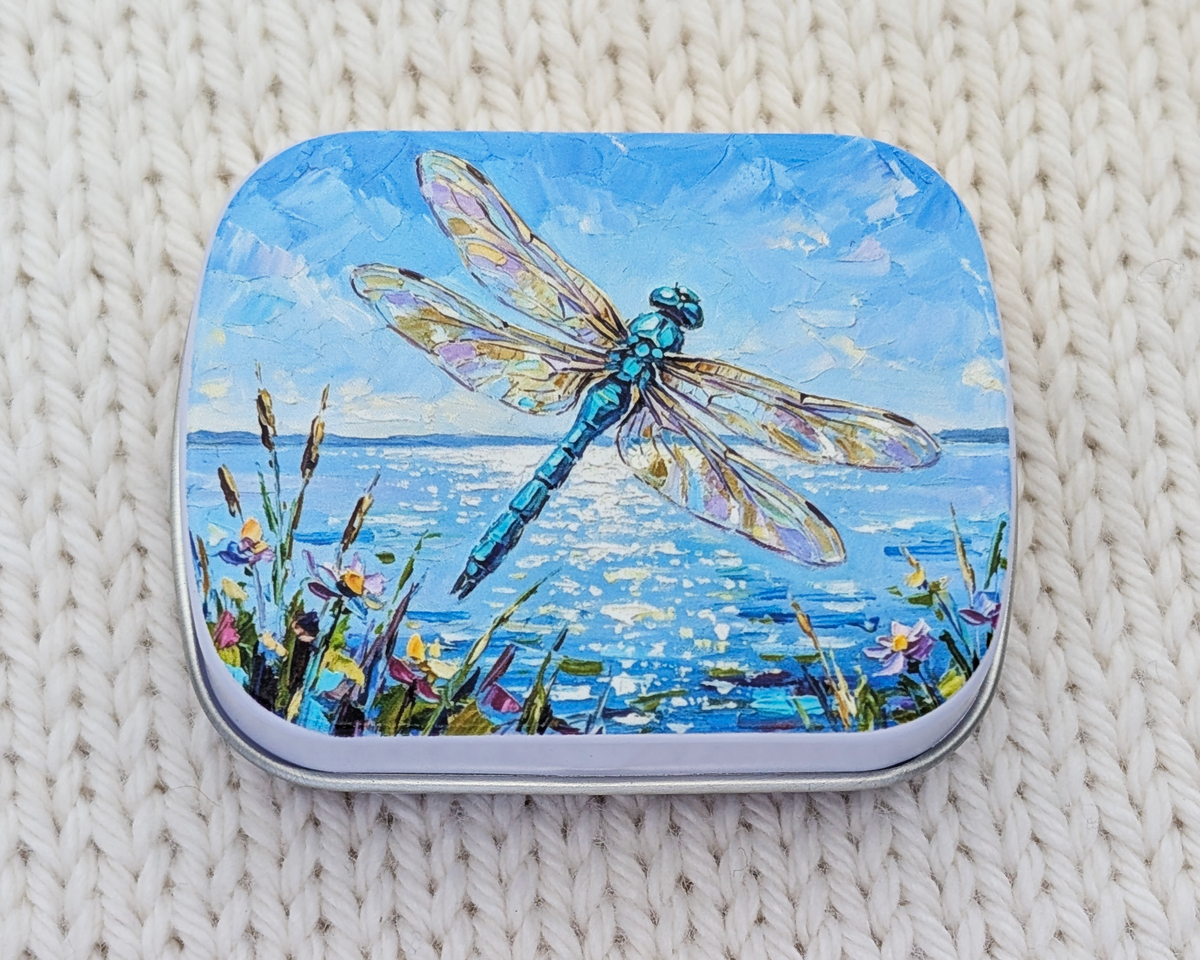 A hinged metal notions tin featuring a colorful painted illustration of a blue dragonfly over water with wildflowers, placed on a cream-colored knitted blanket.