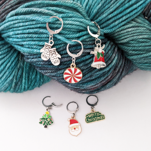 Set of 6 locking crochet style stitch markers shown on worsted weight yarn. Christmas theme includes a Santa, Mittens, Bell, Christmas Tree, Peppermint, and Merry Christmas Sign.