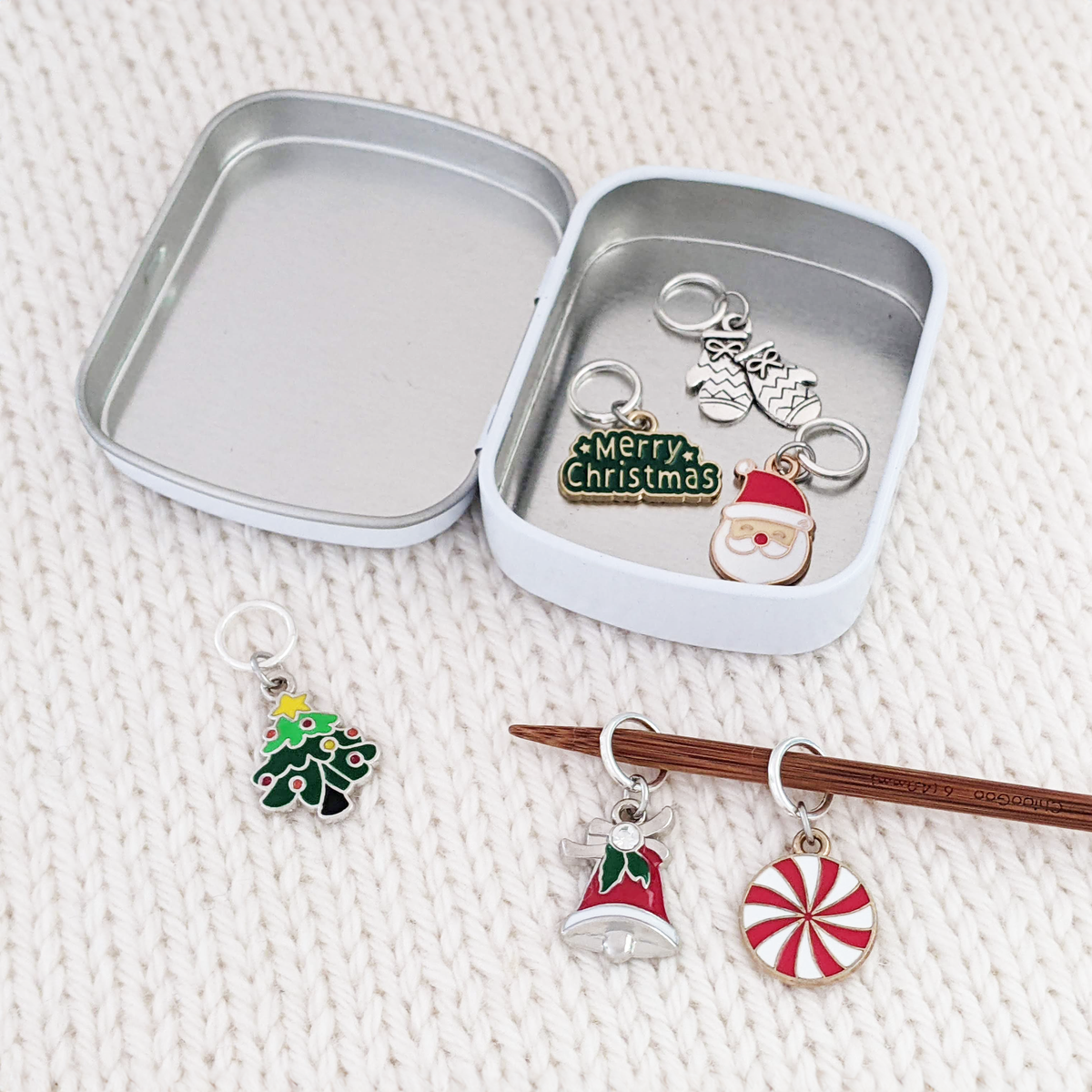 Open metal tin holding Christmas ring stitch markers beside knitting needles