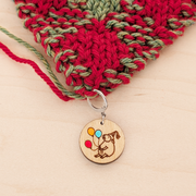 A round birchwood locking stitch marker attached to the edge of red and green double knitted fabric, resting on a light wood surface. The wooden charm features an engraved drawing of a sheep wearing a party hat and holding three hand-painted balloons in red, orange, and blue.