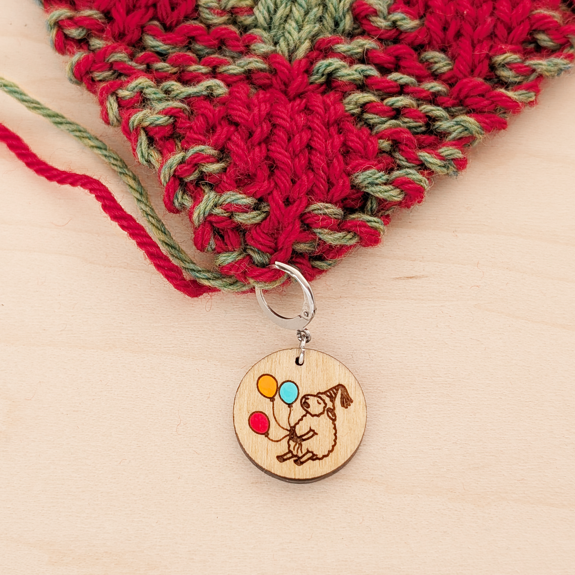 A round birchwood locking stitch marker attached to the edge of red and green double knitted fabric, resting on a light wood surface. The wooden charm features an engraved drawing of a sheep wearing a party hat and holding three hand-painted balloons in red, orange, and blue.