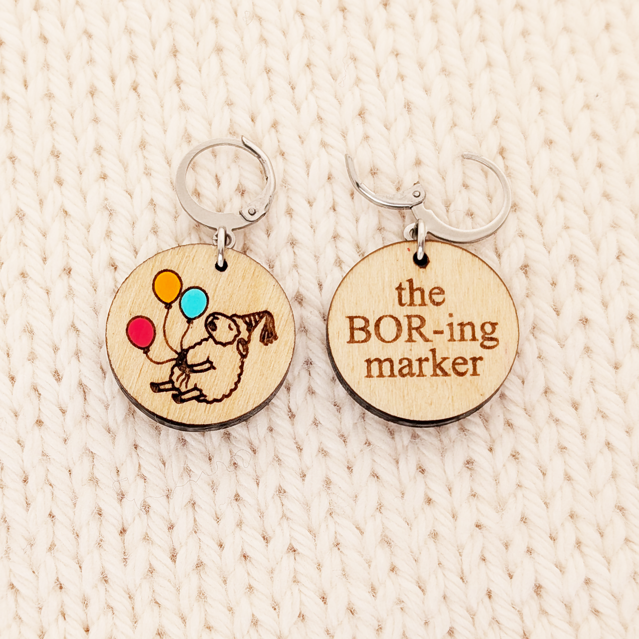 Two round birchwood locking stitch markers resting side-by-side on a textured cream-colored knitted background. The marker on the left shows the front design: an engraved drawing of a sleepy sheep in a party hat, holding three hand-painted balloons in red, orange, and blue. The marker on the right shows the back side of the design: the words 'the BOR-ing marker' engraved in Times New Roman font.