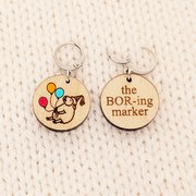 Two round birchwood locking stitch markers resting side-by-side on a textured cream-colored knitted background. The marker on the left shows the front design: an engraved drawing of a sleepy sheep in a party hat, holding three hand-painted balloons in red, orange, and blue. The marker on the right shows the back side of the design: the words 'the BOR-ing marker' engraved in Times New Roman font.