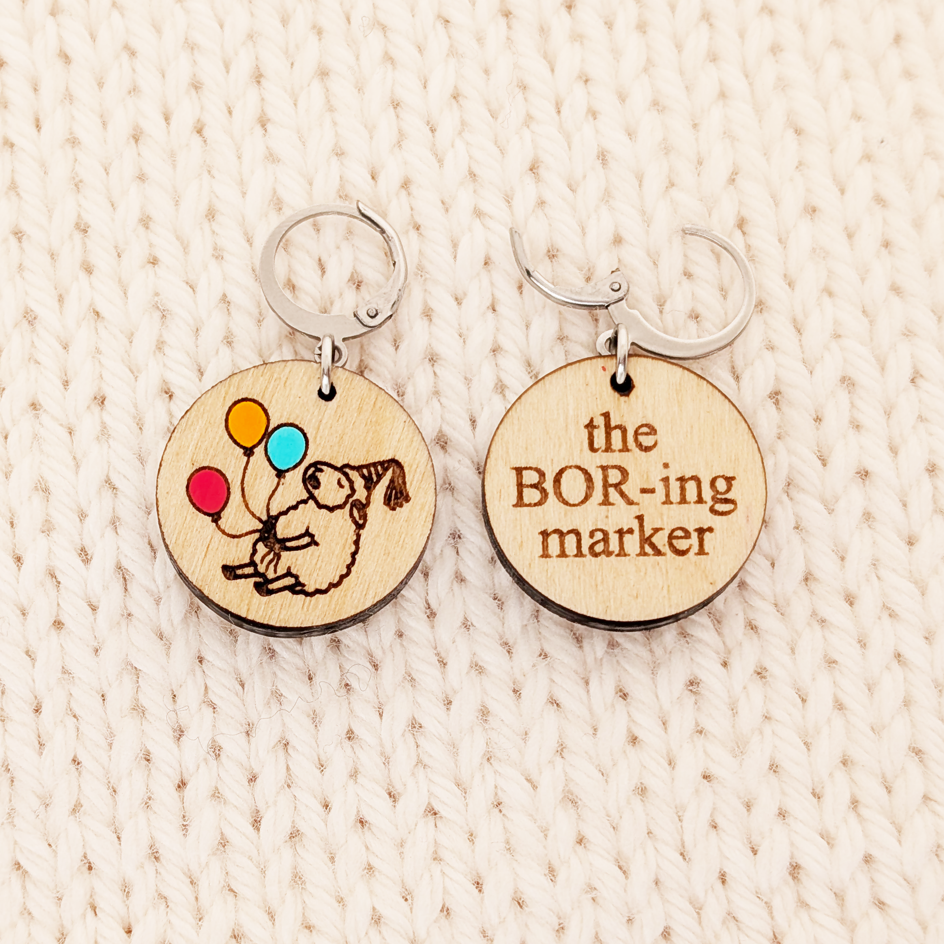 Two round birchwood locking stitch markers resting side-by-side on a textured cream-colored knitted background. The marker on the left shows the front design: an engraved drawing of a sleepy sheep in a party hat, holding three hand-painted balloons in red, orange, and blue. The marker on the right shows the back side of the design: the words 'the BOR-ing marker' engraved in Times New Roman font.