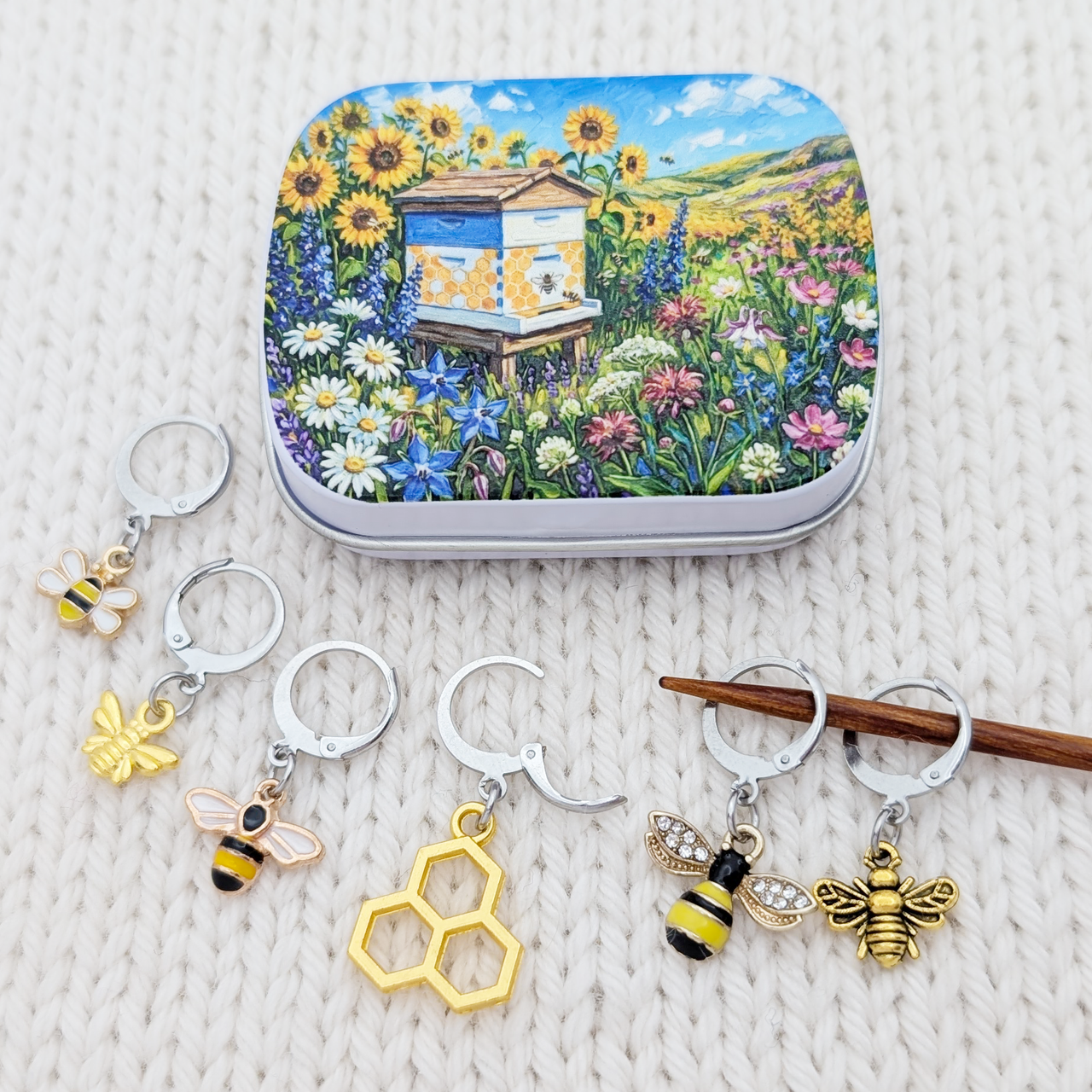 A set of bee-themed crochet and knitting stitch markers and a matching storage tin with a colorful sunflower and beehive illustration, arranged on a white textured knit background.