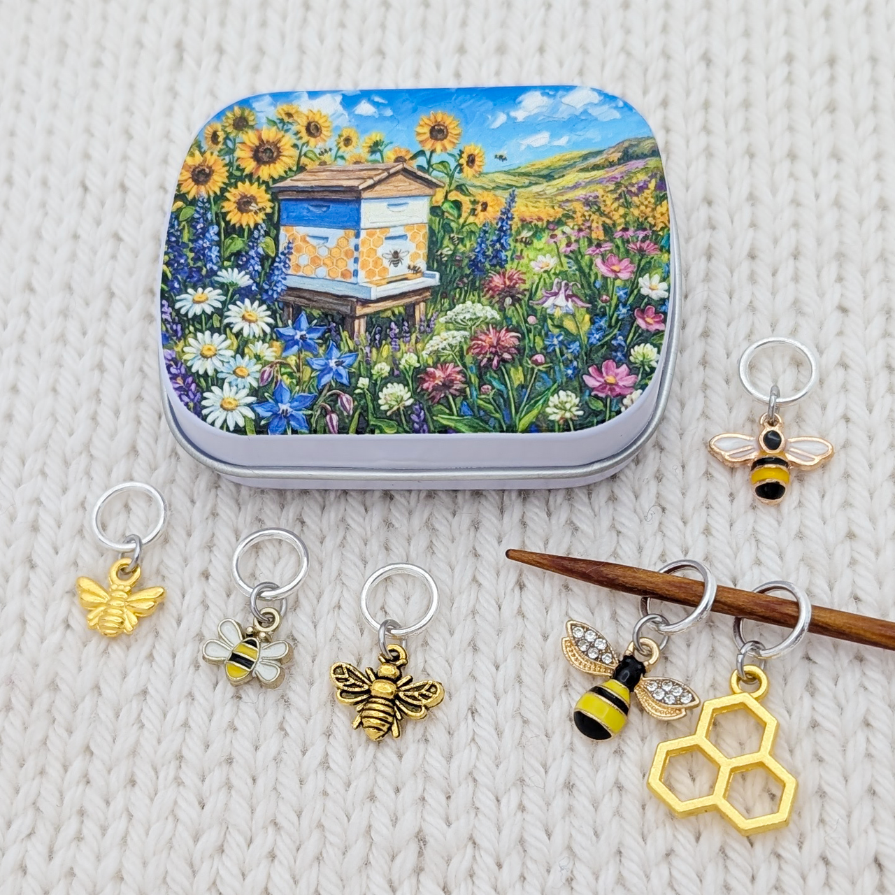 A set of bee-themed stitch markers and a matching tin box, displayed on a cream-colored knitted blanket. The tin box features a colorful illustration of a beehive surrounded by sunflowers and wildflowers.
