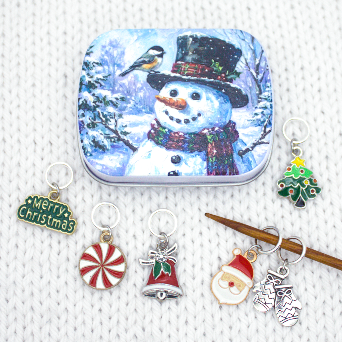 Snowman tin with Christmas stitch markers and wooden knitting needles on a knit swatch