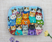 A hinged metal notions tin decorated with a colorful illustration of multiple cats wearing knit sweaters, photographed on a textured beige fabric background with small flowers at the bottom corners.