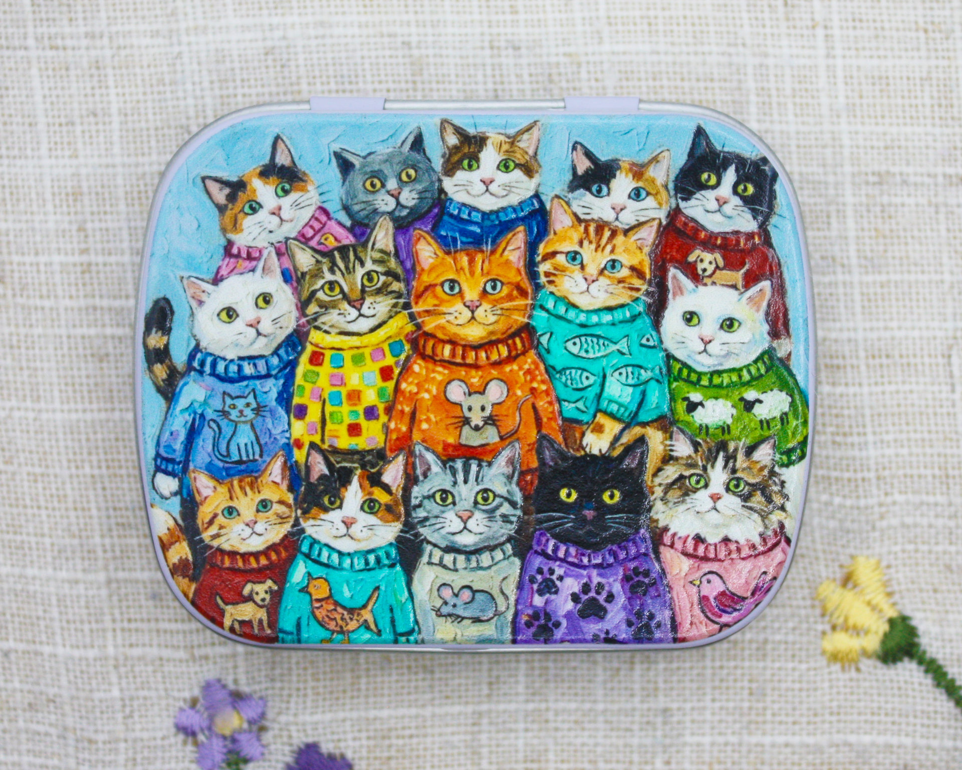 A hinged metal notions tin decorated with a colorful illustration of multiple cats wearing knit sweaters, photographed on a textured beige fabric background with small flowers at the bottom corners.