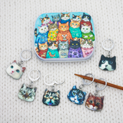 A set of colorful cat-themed locking stitch markers displayed on a cream-colored knit fabric background, with a matching rectangular notions tin featuring a collage of illustrated cats.