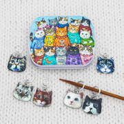 A hinged metal notions tin and six small cat-face knitting stitch markers are arranged on a cream knit sweater. The tin features a colorful illustration of many cats wearing sweaters, and the loose charms show realistic cat faces in different breeds and colors.