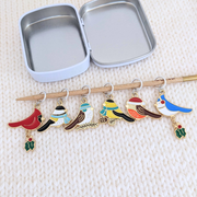Six Christmas bird stitch markers hanging in front of an open tin