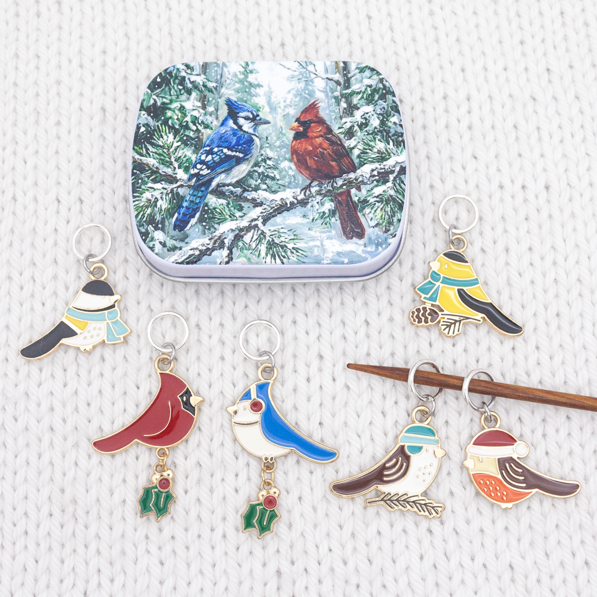 Christmas bird stitch markers beside the tin with wooden knitting needles