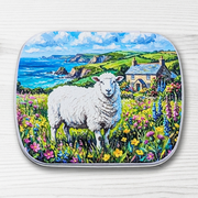 A hinged metal notions tin featuring a colorful landscape illustration of a white sheep in a wildflower field with a cottage and blue sea in the background, shown on a white wood-textured surface.