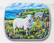 A hinged metal notions tin with a white sheep in a wildflower field with a cottage and blue sea in the background