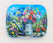 A hinged metal notions tin decorated with a colorful painting of a watering can filled with wildflowers on a light wood background.