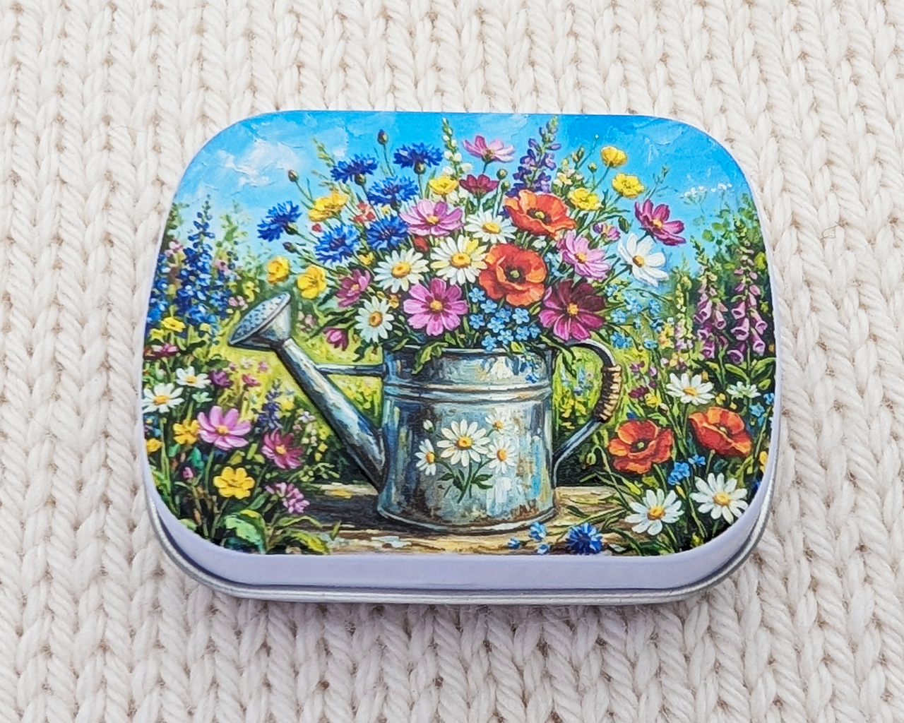 A hinged metal notions tin featuring a colorful illustration of a watering can filled with wildflowers, placed on a cream-colored knitted blanket.