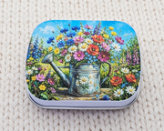 A hinged metal notions tin featuring a colorful illustration of a watering can filled with wildflowers, placed on a cream-colored knitted blanket.