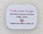 The bottom side of a hinged metal notions tin printed with the brand name 'Pretty Warm Designs' and 'Knitting & Crochet Notions, Cottage Garden' in pink and black lettering, with a small pink rose illustration and Designed and crafted in Victoria BC. The tin is placed on a cream-colored knitted blanket background.