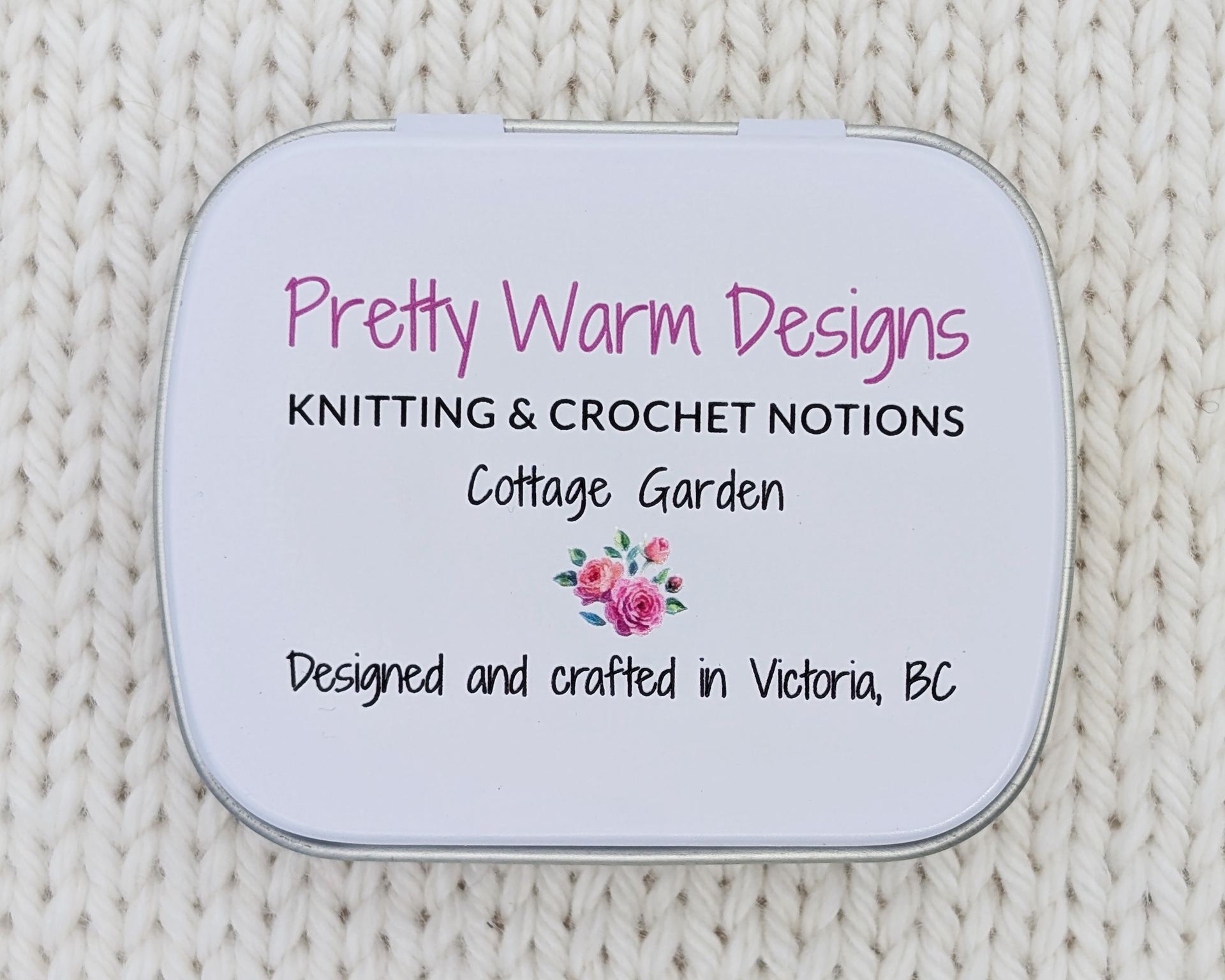 The bottom side of a hinged metal notions tin printed with the brand name 'Pretty Warm Designs' and 'Knitting & Crochet Notions, Cottage Garden' in pink and black lettering, with a small pink rose illustration and Designed and crafted in Victoria BC. The tin is placed on a cream-colored knitted blanket background.