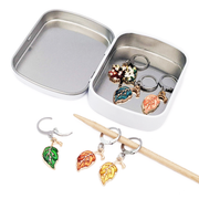 Set of 6 locking stitch markers with glossy enamel metal charms in the shape of leaves and a tree, shown with a white storage tin. Handmade by Pretty Warm Designs.