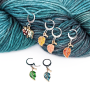 Set of 6 locking stitch markers with glossy enamel metal charms in the shape of leaves and a tree, shown on a skein of worsted weight yarn. Handmade by Pretty Warm Designs.