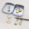 Set of six white, yellow and green enamel metal daisy charms with lever back clasps stitch markers for crochet and decorative tin with logo by Pretty Warm Designs, shown on a 4mm knitting needle