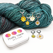 Set of six white, yellow and green enamel metal daisy charms with lever back clasps stitch markers for crochet and decorative tin with logo by Pretty Warm Designs, shown with a Malabrigo worsted weight yarn skein