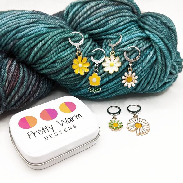 Set of six white, yellow and green enamel metal daisy charms with lever back clasps stitch markers for crochet and decorative tin with logo by Pretty Warm Designs, shown with a Malabrigo worsted weight yarn skein