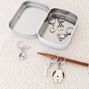 Set of 6 pet dog themed locking stitch markers for crochet with decorative storage tin. Includes a love my dog heart, a bowl with a bone, a black and white beagle, a bone, a paw, and an I love dogs sign.  Shown with a knitting needle.
