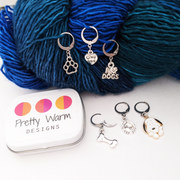 Set of 6 pet dog themed locking stitch markers for crochet. Includes a love my dog heart, a bowl with a bone, a black and white beagle, a bone, a paw, and an I love dogs sign. Storage tin included. Shown with worsted weight yarn.