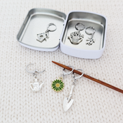 Set of one green and yellow enamel daisy and five antiqued silver alloy gardening themed locking stitch markers for crochet and decorative storage tin with logo by Pretty Warm Designs Inc, shown on a knitting needle