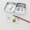 Set of one green and yellow enamel daisy and five antiqued silver alloy gardening themed locking stitch markers for crochet and decorative storage tin with logo by Pretty Warm Designs Inc, shown on a knitting needle