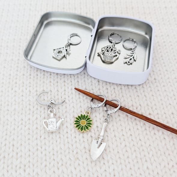 Set of one green and yellow enamel daisy and five antiqued silver alloy gardening themed locking stitch markers for crochet and decorative storage tin with logo by Pretty Warm Designs Inc, shown on a knitting needle
