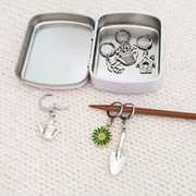 Set of one green and yellow enamel daisy and five antiqued silver alloy gardening themed locking stitch markers shown with a decorative white storage tin and displayed on a knitting needle, by Pretty Warm Designs Inc