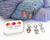 Set of six multicolored enamel on silver toned alloy owl locking stitch markers for crochet with decorative tin by Pretty Warm Designs, shown on worsted weight yarn