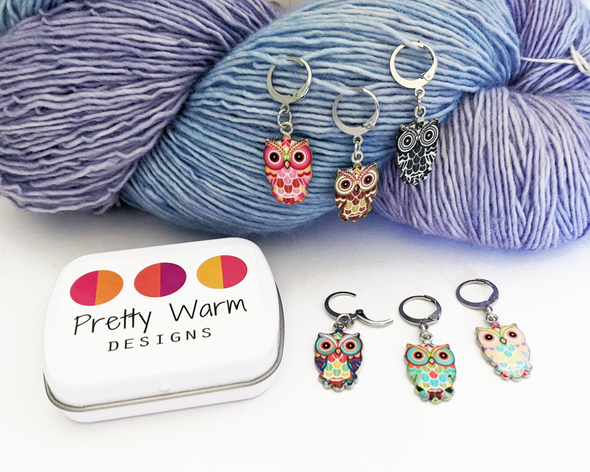 Set of six multicolored enamel on silver toned alloy owl locking stitch markers for crochet with decorative tin by Pretty Warm Designs, shown on worsted weight yarn