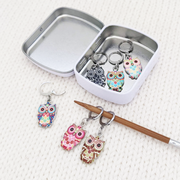 Set of six multicolored enamel on silver toned alloy owl locking stitch markers for crochet with decorative tin by Pretty Warm Designs, shown with knitting needle