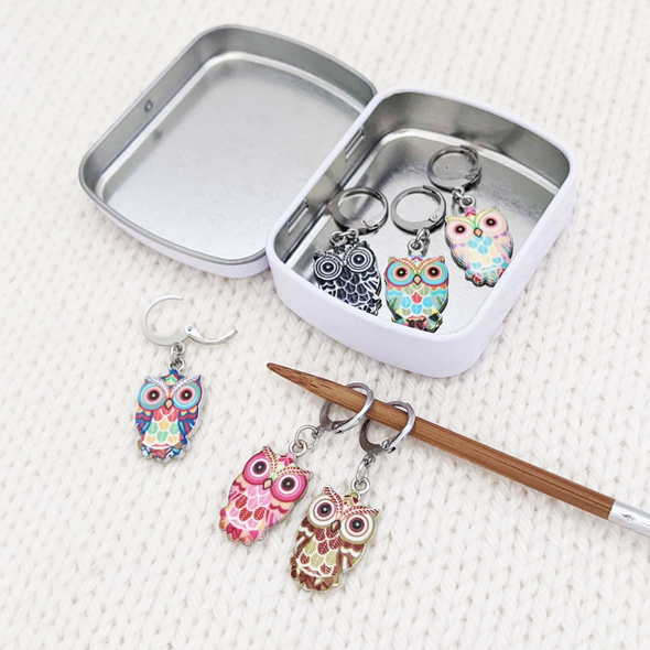 Set of six multicolored enamel on silver toned alloy owl locking stitch markers for crochet with decorative tin by Pretty Warm Designs, shown with knitting needle