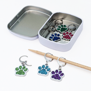 Pretty Warm Designs Sparkle Paws locking stitch markers for knitting and crochet. Three glitter enamel paw print charms sit inside an open rectangular hinged notions tin. Outside the tin, two markers are hooked onto a wooden knitting needle and one lies open on a white surface. Features 316 stainless steel leverback locking clasps.