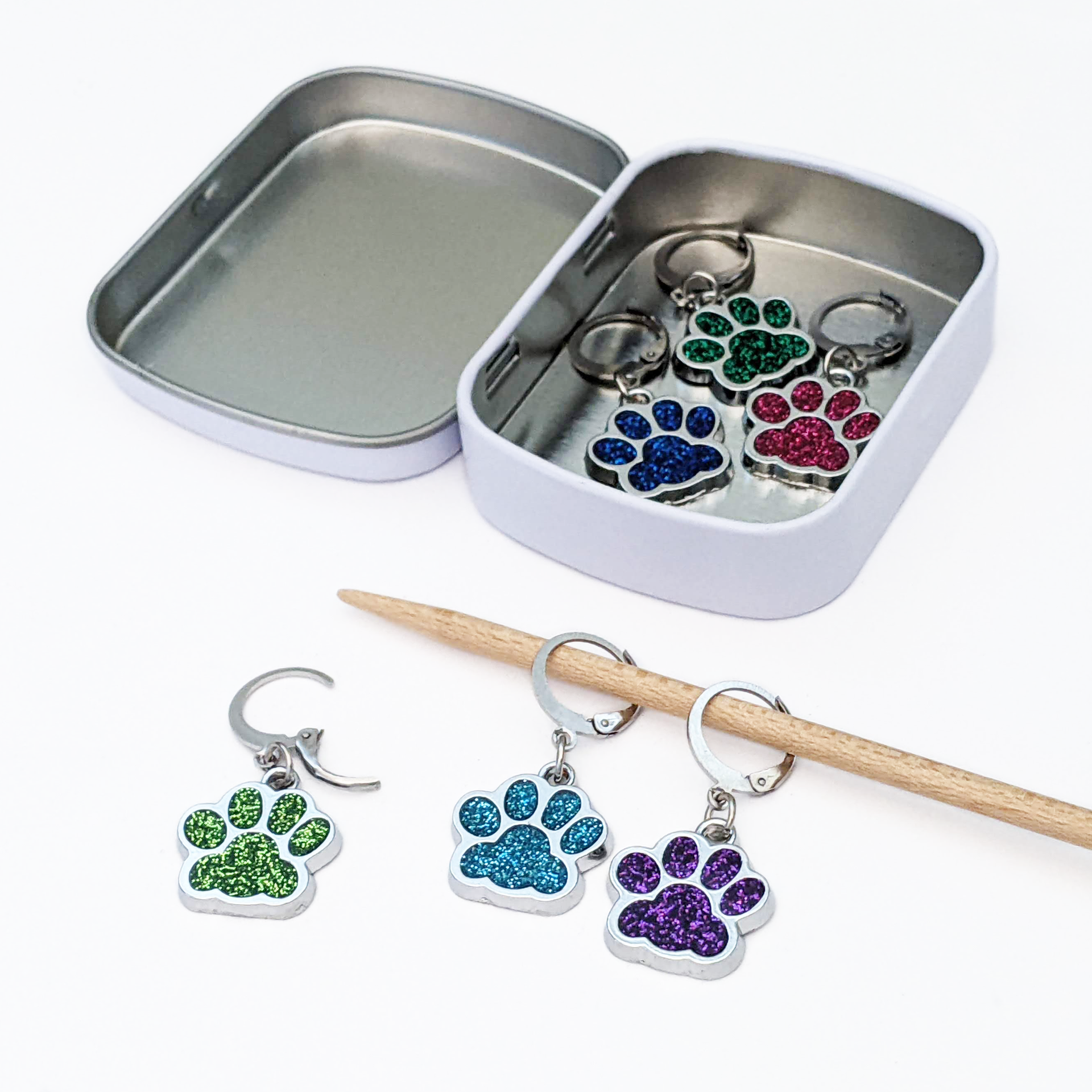 Pretty Warm Designs Sparkle Paws locking stitch markers for knitting and crochet. Three glitter enamel paw print charms sit inside an open rectangular hinged notions tin. Outside the tin, two markers are hooked onto a wooden knitting needle and one lies open on a white surface. Features 316 stainless steel leverback locking clasps.