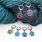 Sparkle Paws locking stitch markers for knitting and crochet by Pretty Warm Designs. Two glitter enamel paw print charms in magenta and purple are hooked into a skein of variegated yarn, while four markers in shades of blue and green lie flat on a white surface. Features 316 stainless steel leverback clasps.