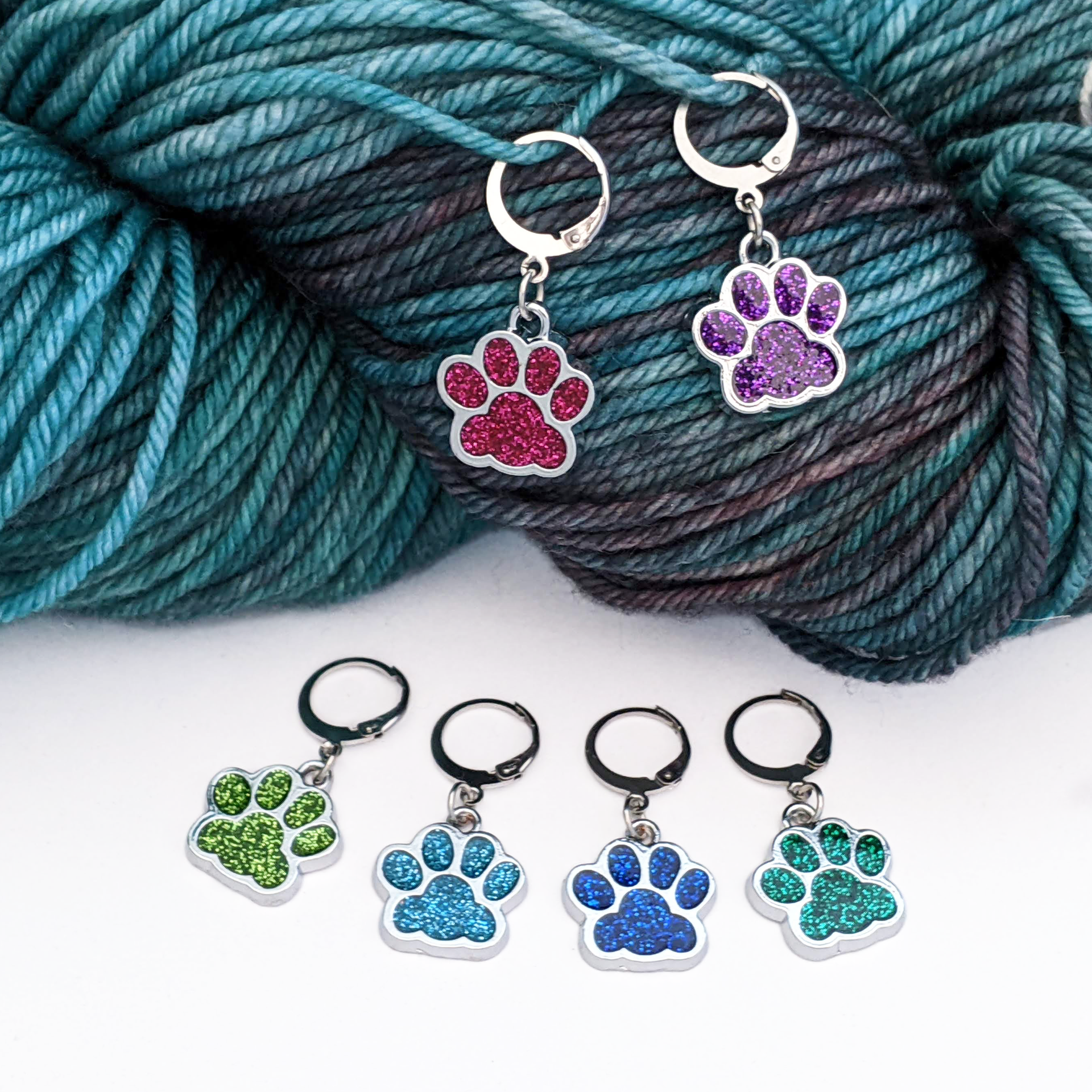 Sparkle Paws locking stitch markers for knitting and crochet by Pretty Warm Designs. Two glitter enamel paw print charms in magenta and purple are hooked into a skein of variegated yarn, while four markers in shades of blue and green lie flat on a white surface. Features 316 stainless steel leverback clasps.