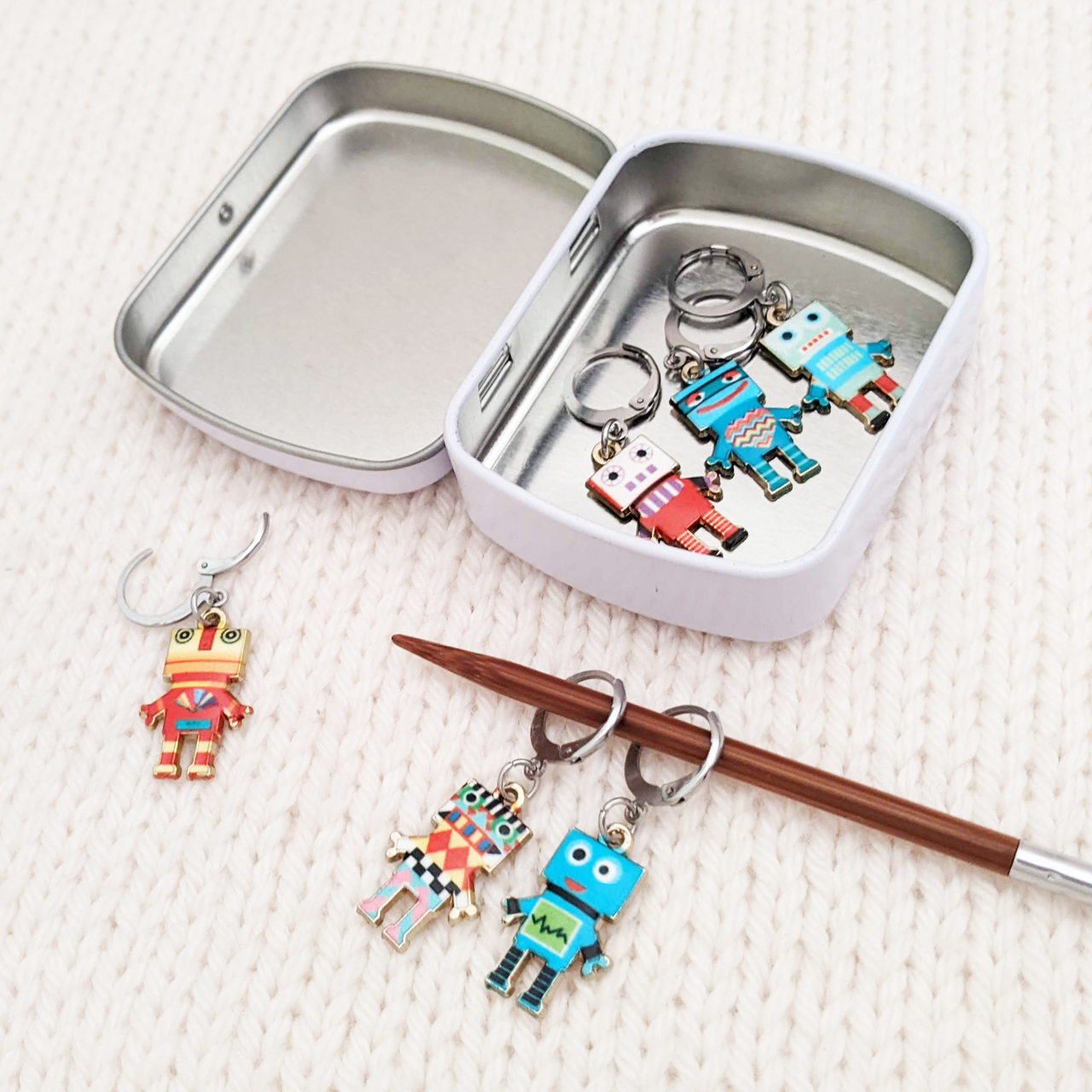 Set of 6 Removable stitch markers for crochet or knitting with multicoloured robot charms and storage tin. Shown on a knitting needle with opening clasp.