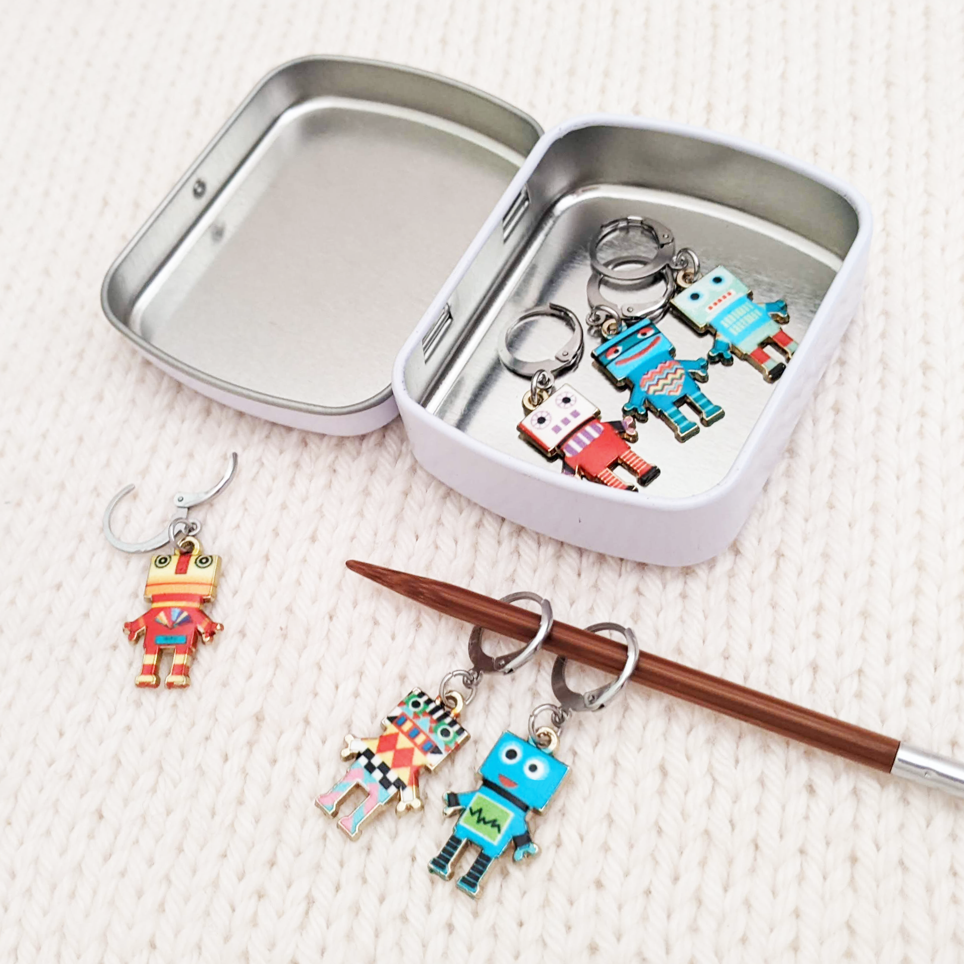 Set of 6 Removable stitch markers for crochet or knitting with multicoloured robot charms and storage tin. Shown on a knitting needle with opening clasp.