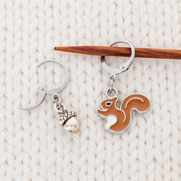 A close-up of the enamel squirrel and silver acorn locking stitch markers. One hangs securely from a wooden knitting needle and the other rests on cream colored knitting.